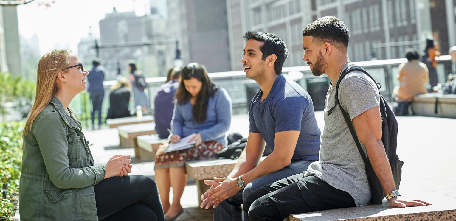 Three seated people engaging in lively conversation under a blooming cherry blossom tree. On the left, a woman with shoulder-length blonde hair wears glasses, a green jacket, and black pants. Her hands are clasped in her lap. In the middle, a man with short black hair wears a blue collared shirt and blue jeans. His widespread hands gesture downward. On the right, a man with cropped black hair and a beard wears a gray t-shirt, a black backpack, and black pants. He leans back on his hands.